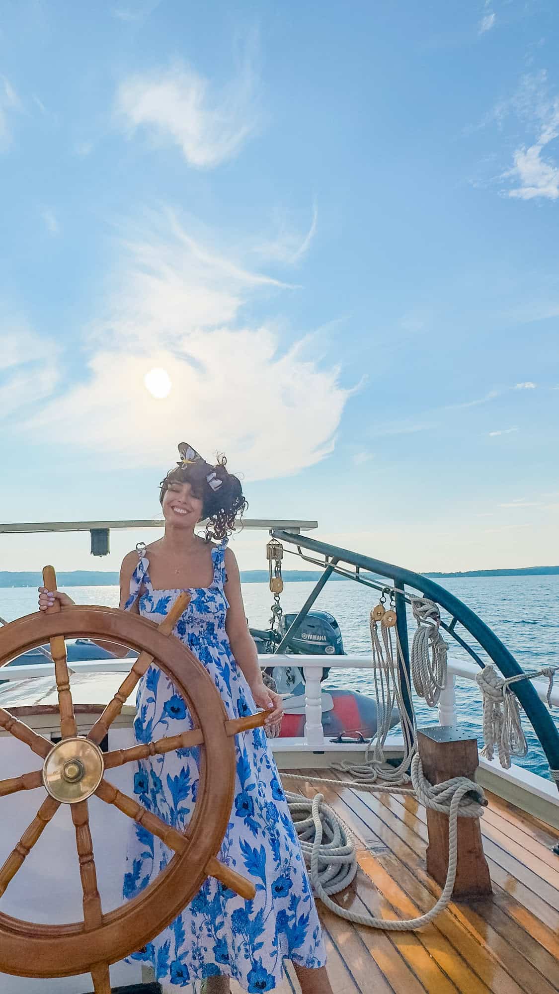 Woman in blue dress standing at a ship's wheel on a sunny day with clear skies.