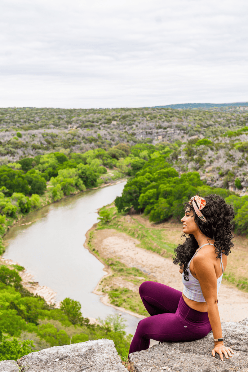 Person sitting on a cliff overlooking a river and green landscape.