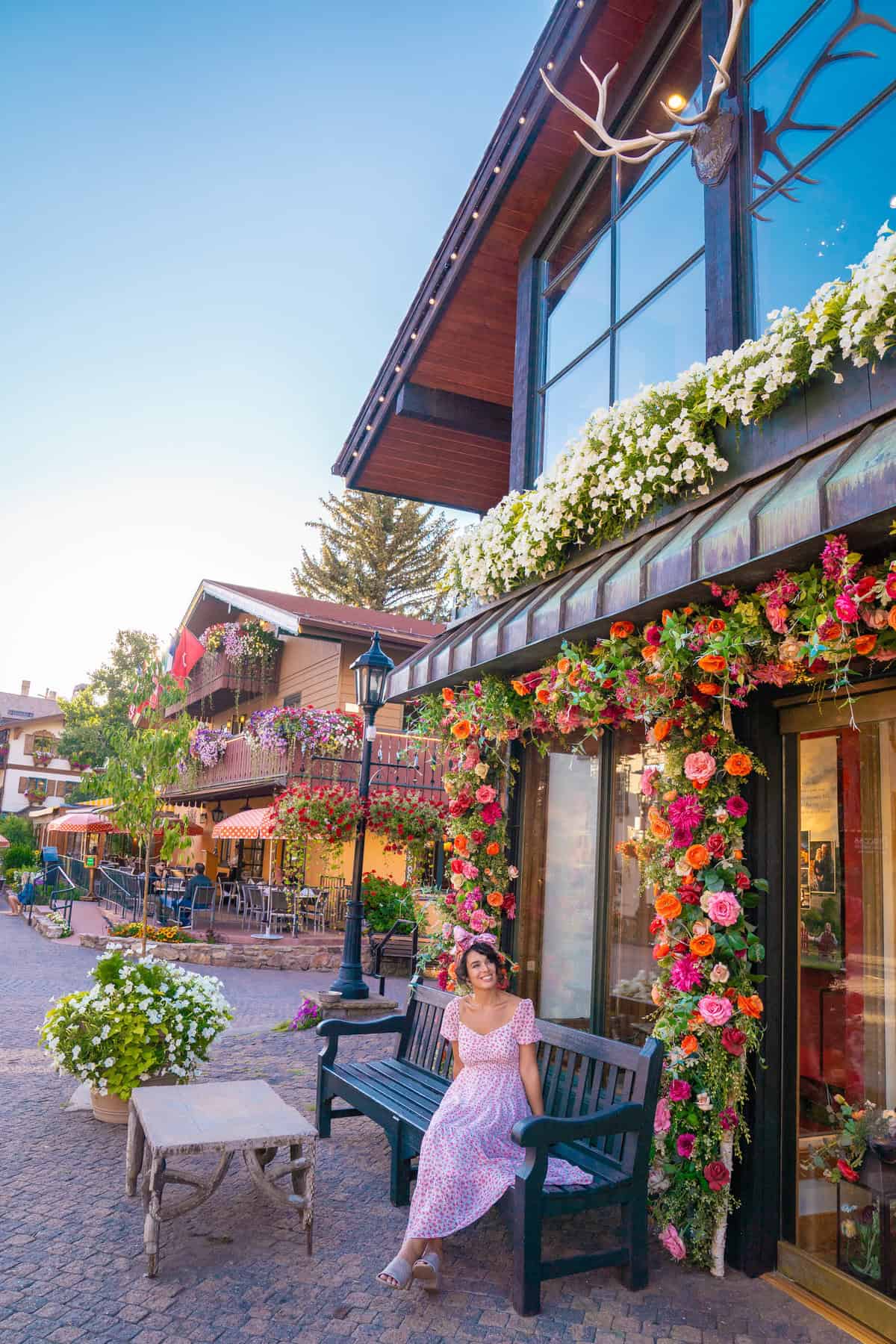 A person in a pink dress sits on a bench by a flower-adorned shopfront