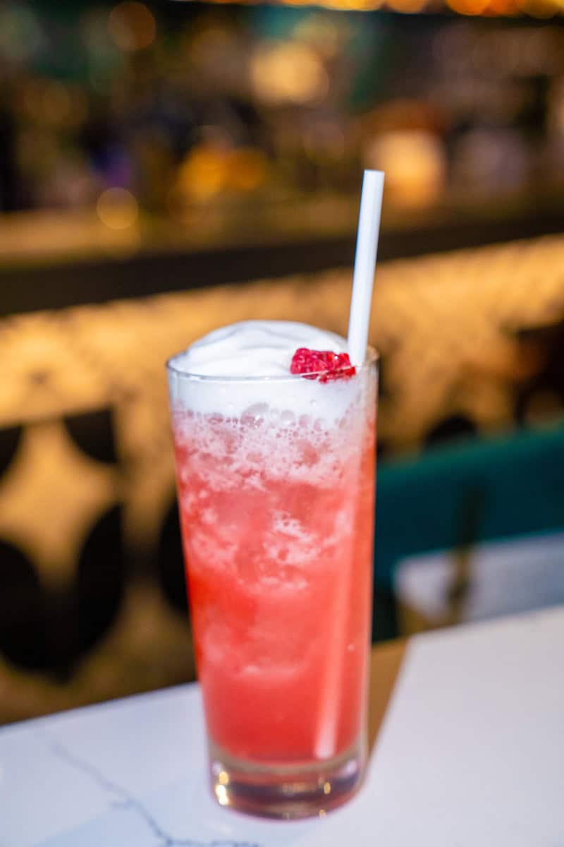 A frothy red cocktail with a white straw and garnish on a bar counter.