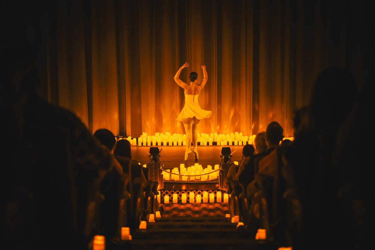 Ballerina performing on stage, audience in silhouette, warm candlelit setting.