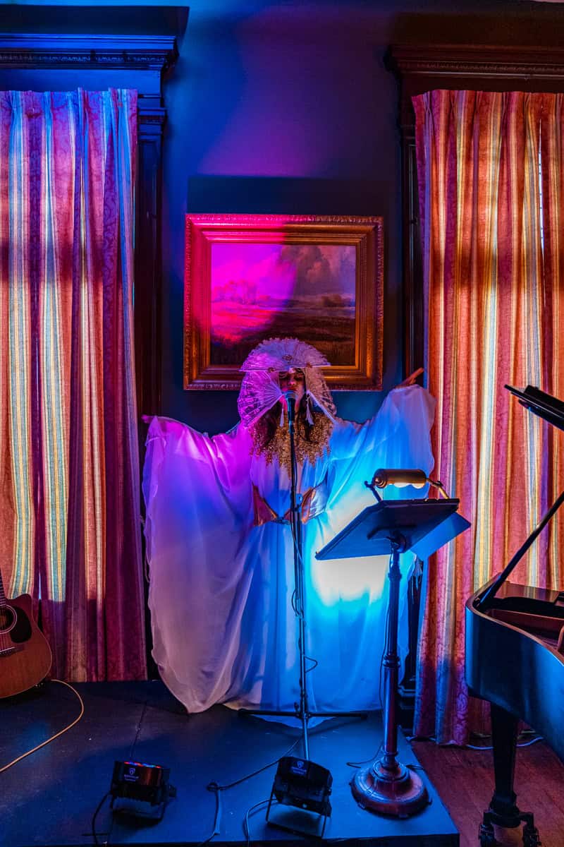 A woman in a white dress and a mask gracefully sings in front of the the piano