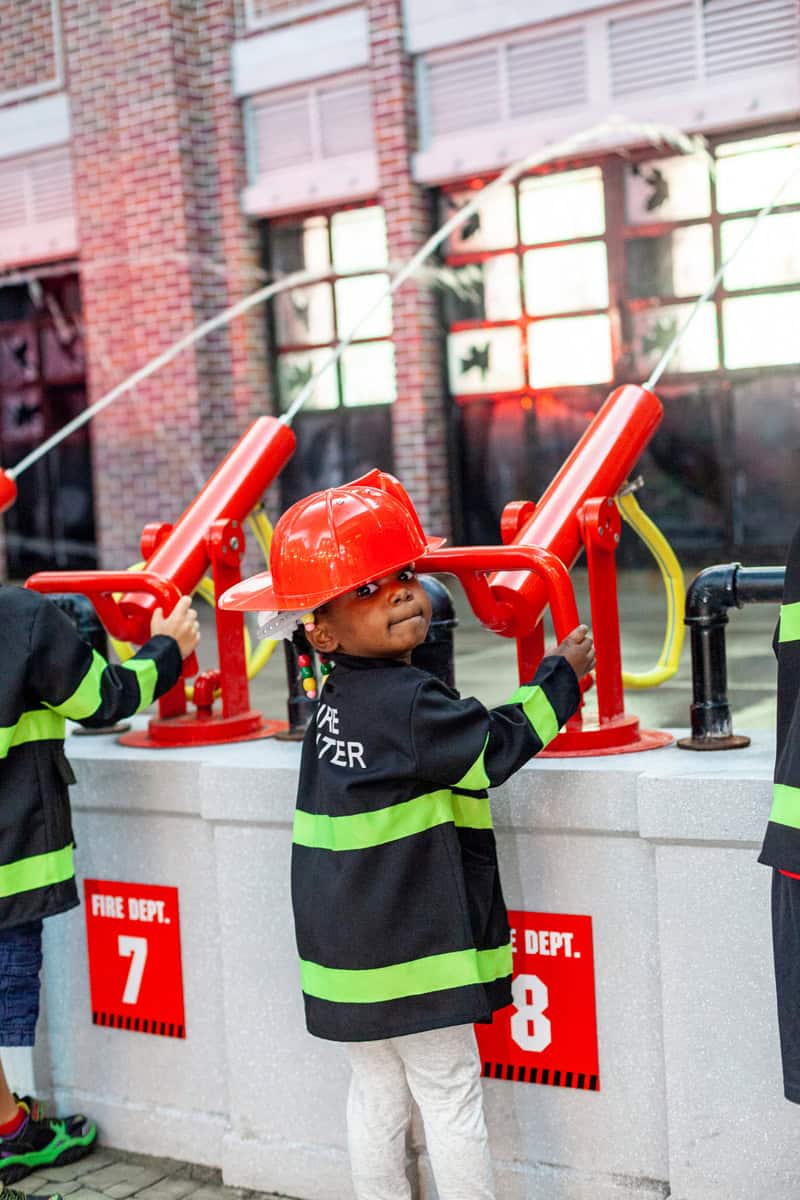 Children in firefighter jackets playing with water at a fire station exhibit.