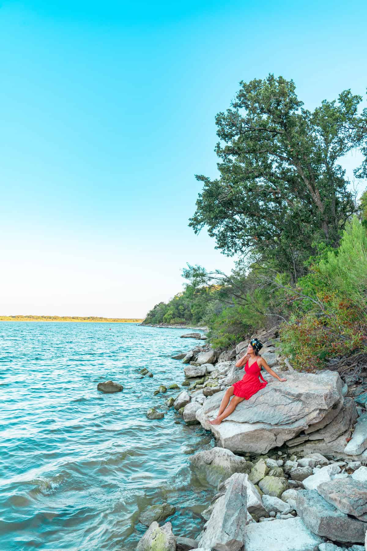 A woman wearing a red dress relaxes on a rock near the wate