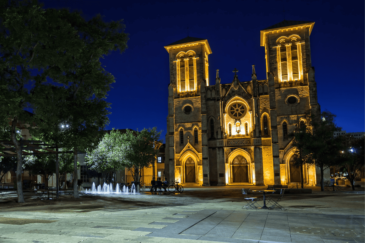 Illuminated Gothic church at night with surrounding trees and fountain.