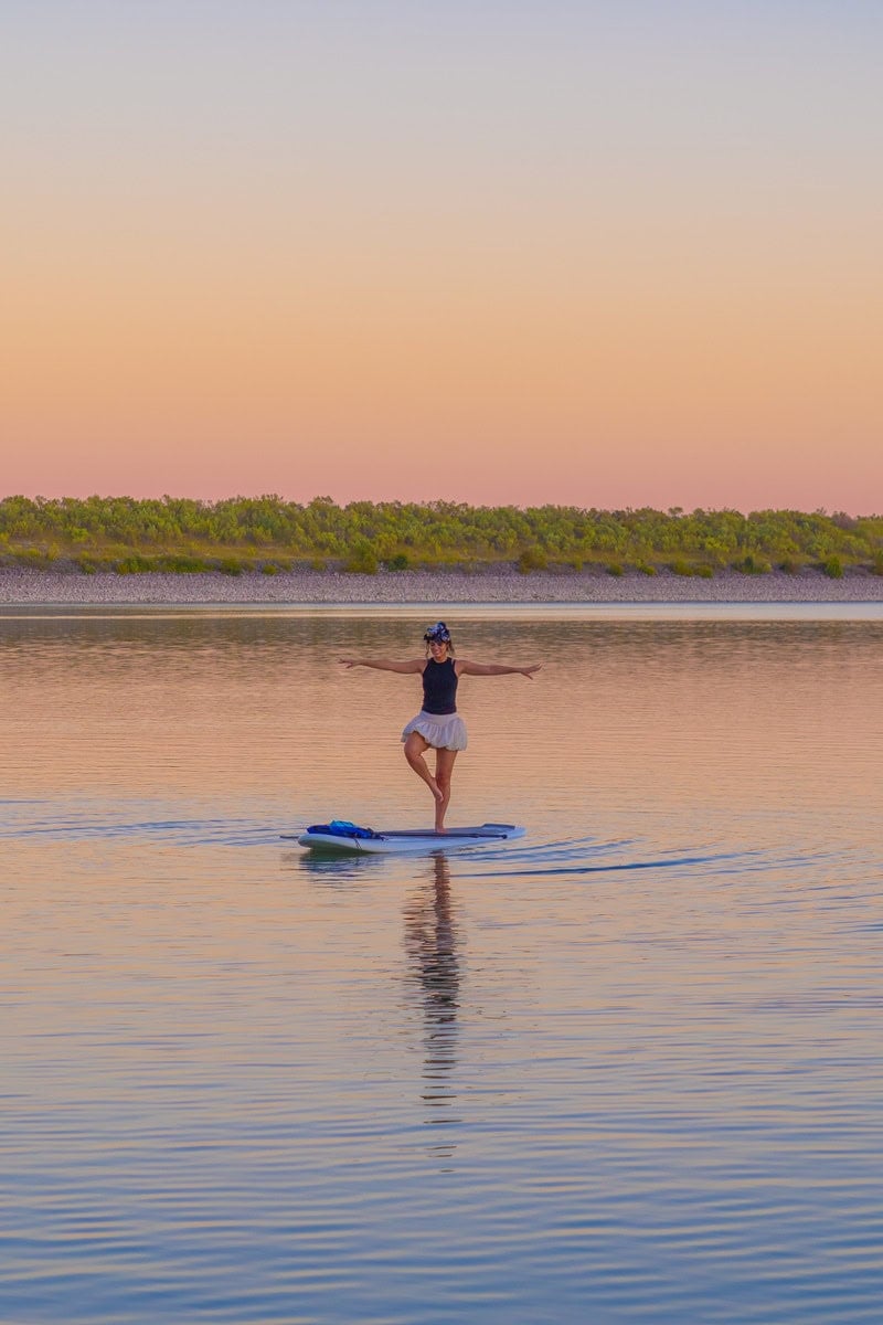 A person balancing on a paddleboard on calm water at sunset.