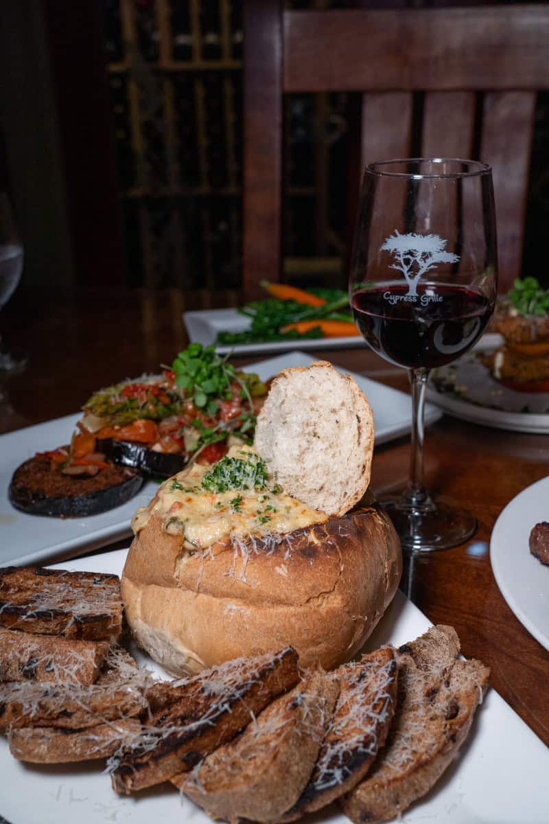 Bread bowl with creamy soup, sliced baguette, wine glass, and side dishes.