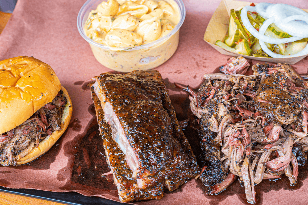 BBQ meal with ribs, pulled pork sandwich, potato salad, and pickles.