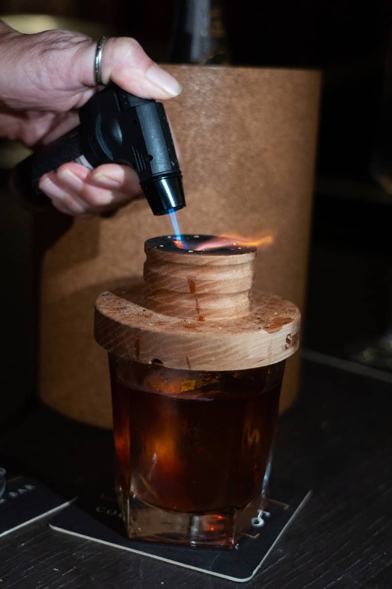 Hand using a blowtorch on a wooden garnish atop a cocktail glass.