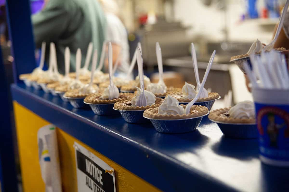 Rows of pies with whipped cream and plastic forks on a counter.