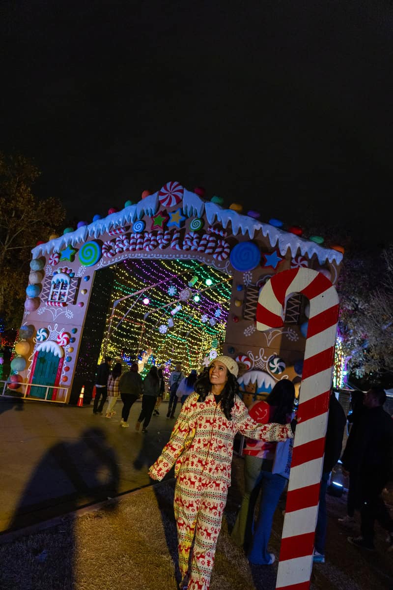 woman in pajamas smiles while standing in front of a colorful gingerbread house