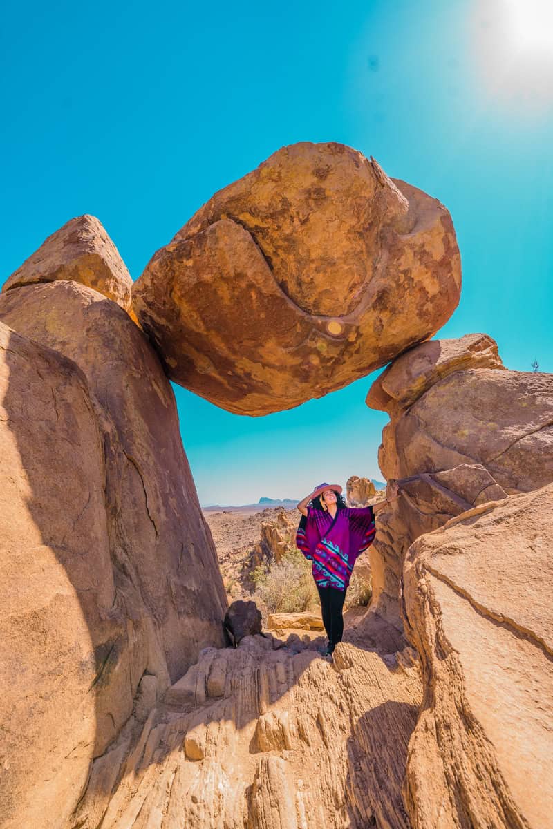 Person standing below a precariously balanced rock in a desert landscape.