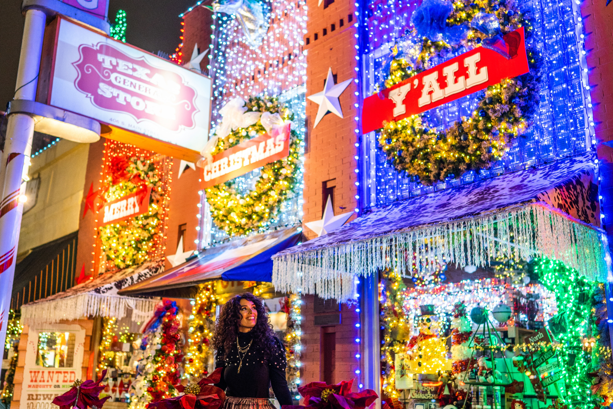 A cheerful woman poses in front of a store sparkling with Christmas lights, 