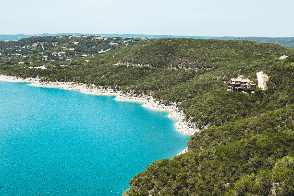 Aerial view of a turquoise lake beside a forested hill with houses.