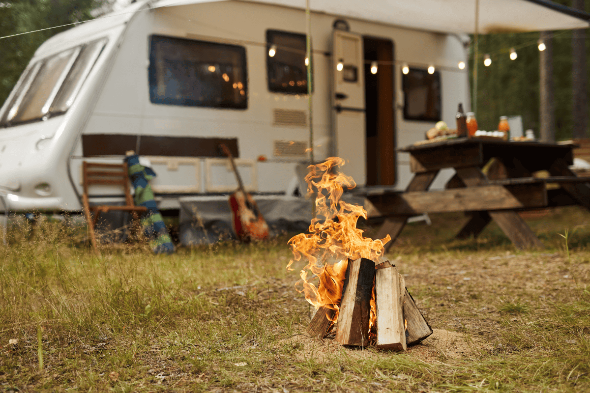 Campfire in focus with a caravan and picnic table in the background.