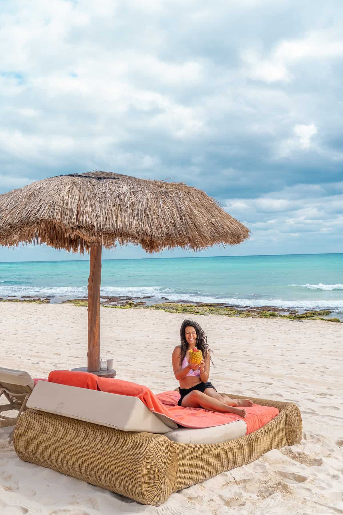 A person relaxes on a wicker lounge chair under a thatched umbrella