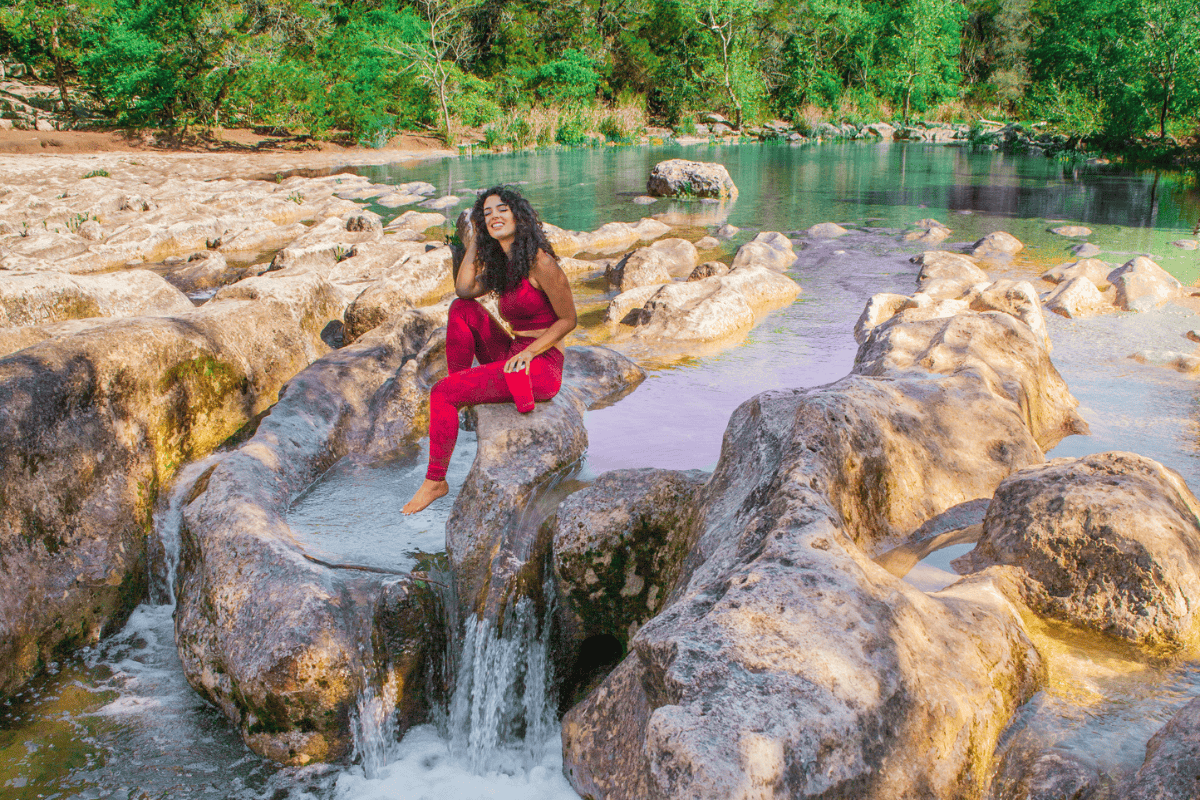 A person sits on a smooth rock by a tranquil stream