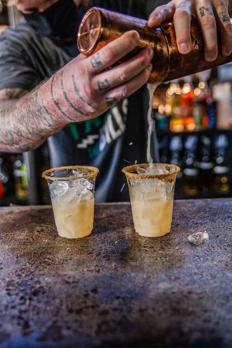 A bartender with tattooed arms pours a cocktail from a shaker