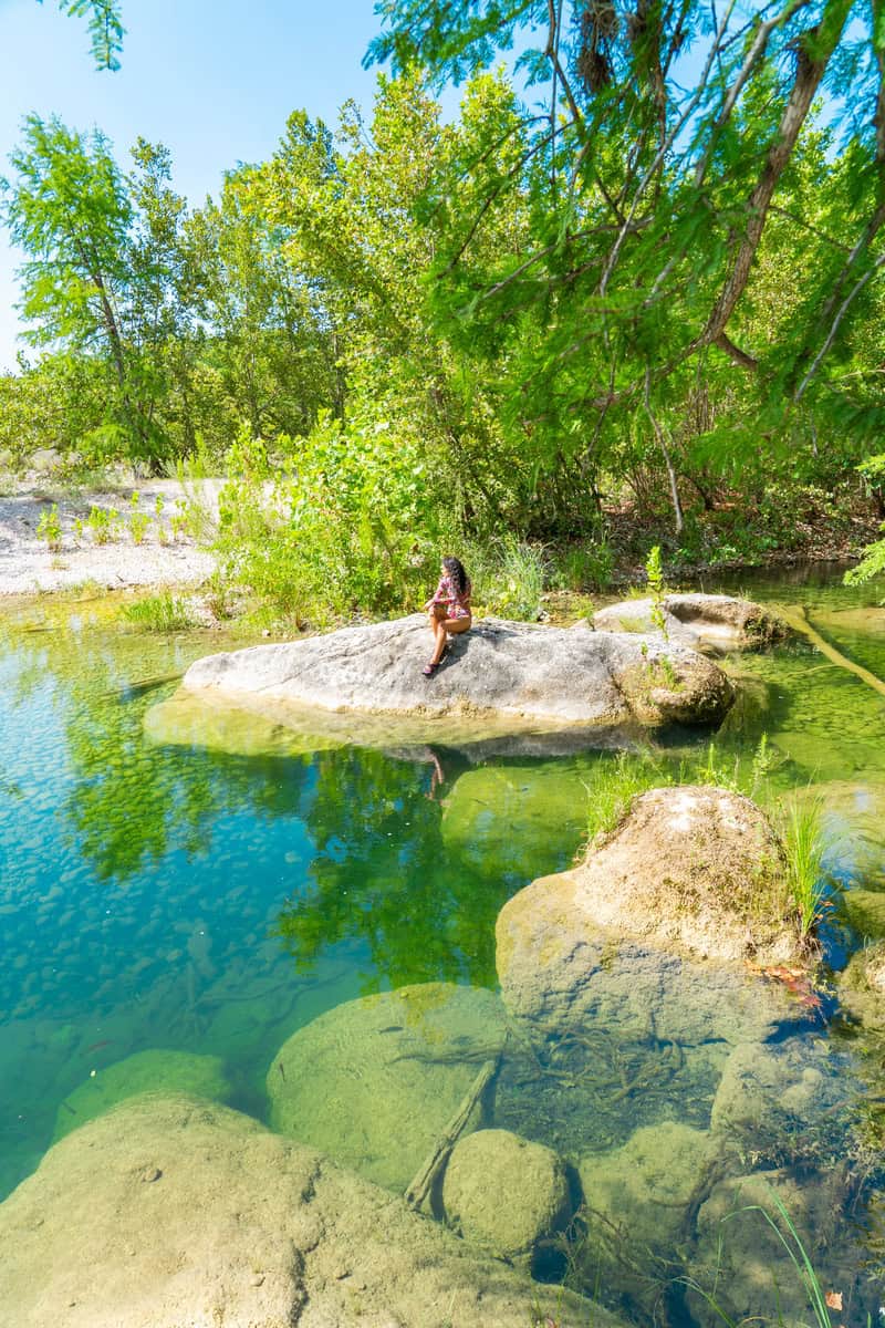 A girl sits on a large rock by a serene, clear river, surrounded by greenery