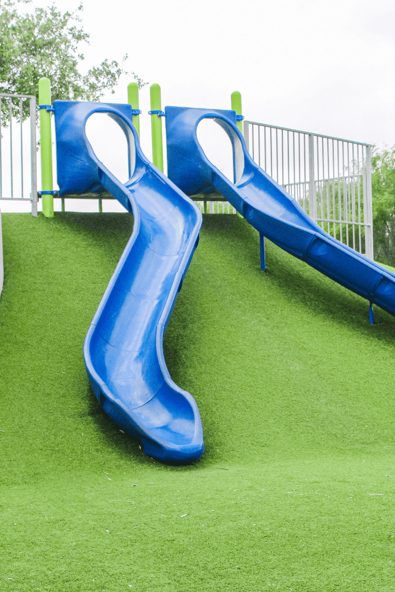 Two bright blue playground slides descend from a grassy hill