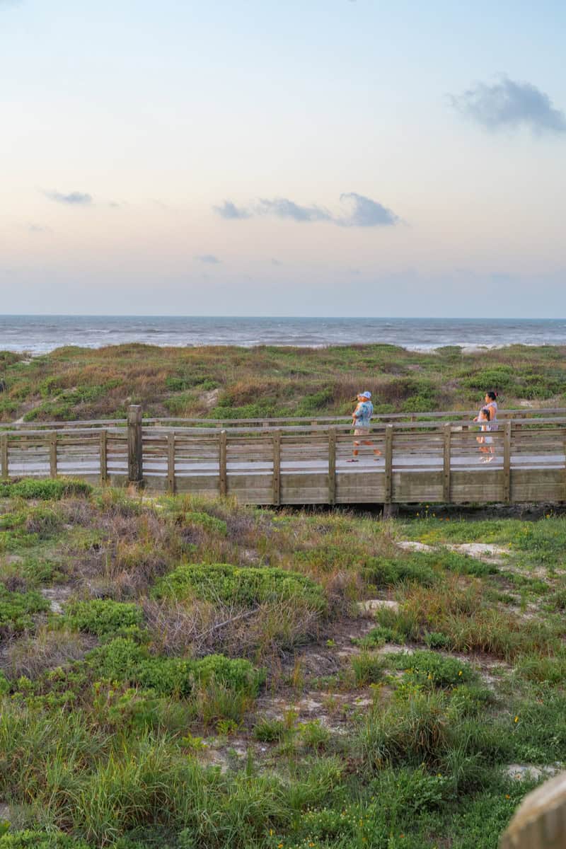 Two people stroll on a wooden boardwalk along a beach at sunset