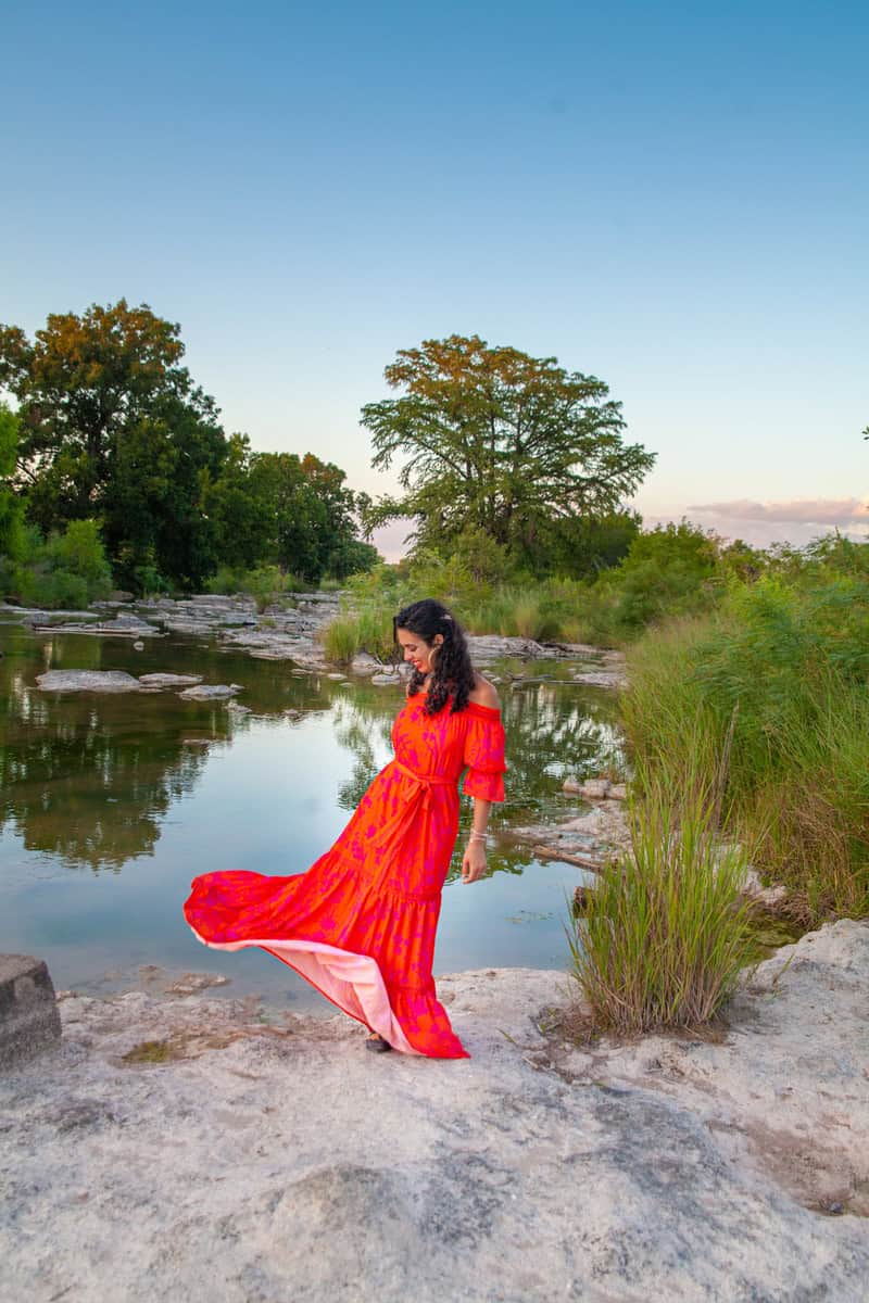 A woman in a vibrant red dress stands thoughtfully by a calm river