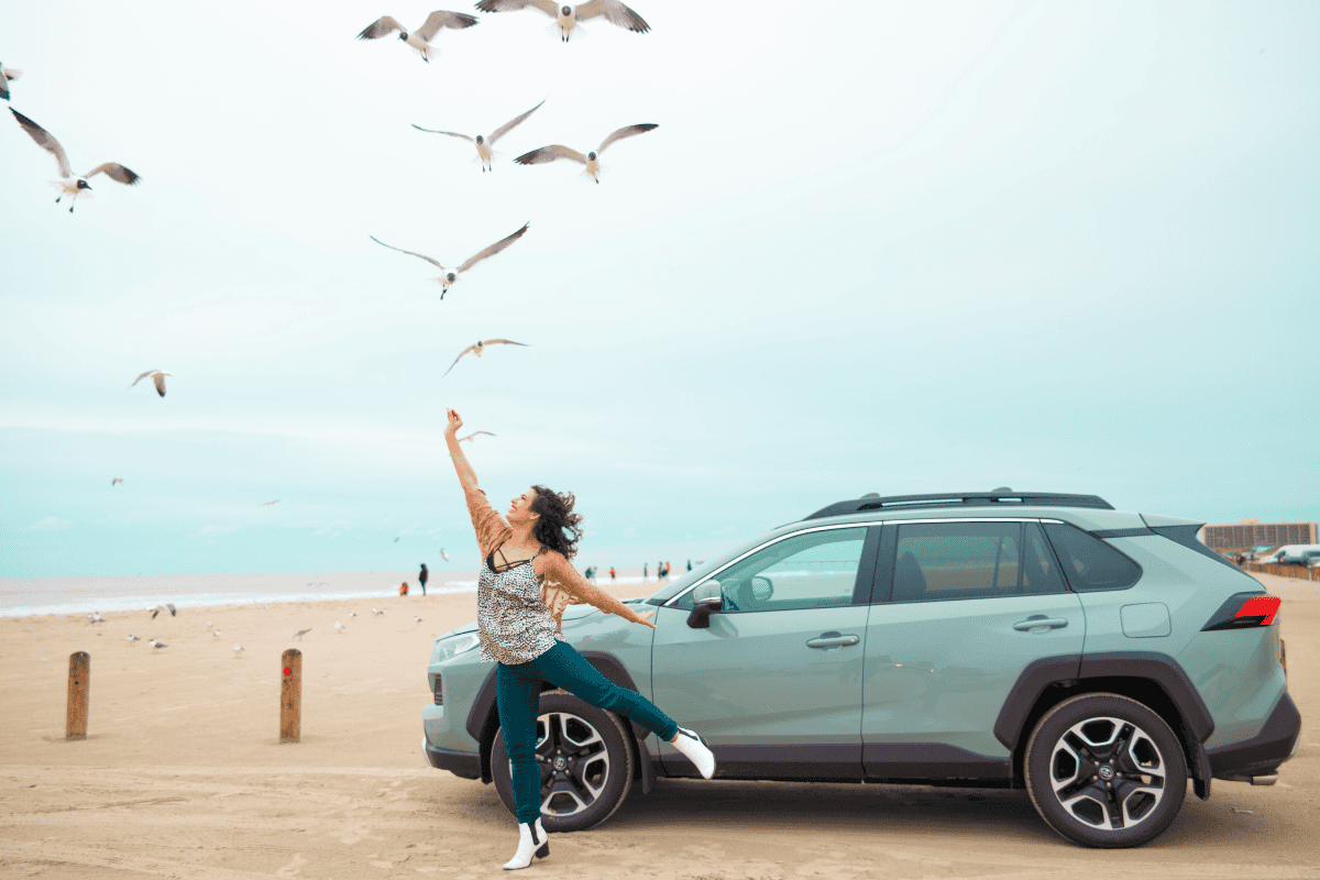 A woman joyfully dances beside a green car on a sandy beach