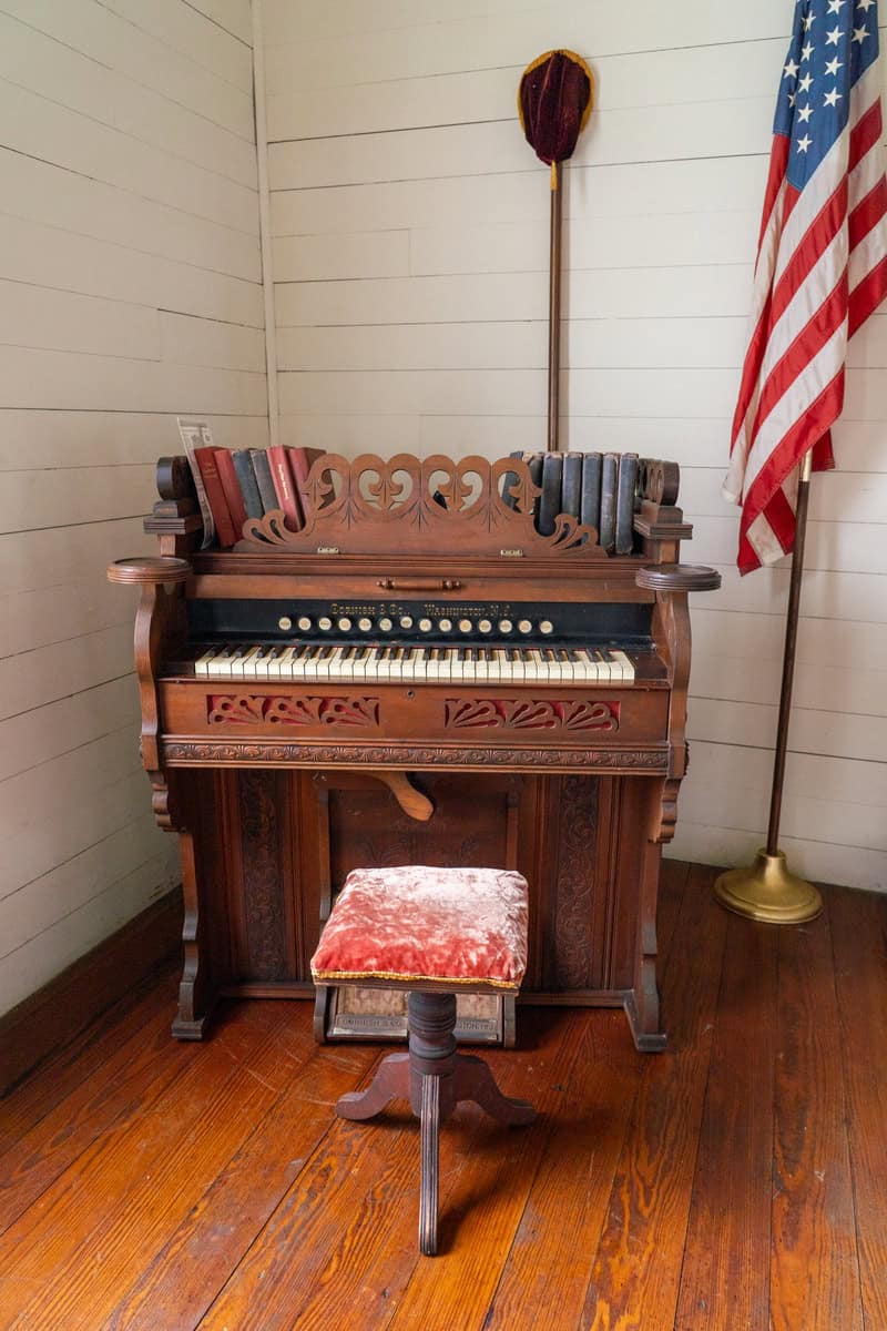 Antique organ, displaying books on top, alongside a plush velvet stool and an American flag inside the chapel of Tomball Museum Center