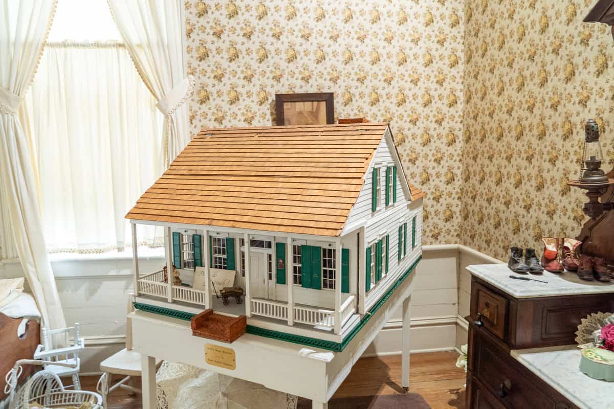 A vintage dollhouse with green shutters and a wooden roof, displayed in a room at Tomball Museum Center