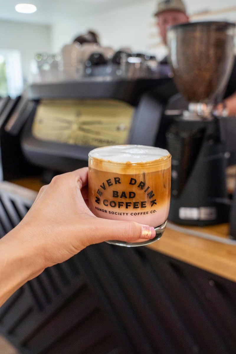 a hand holds a glass of creamy layered coffee with a slogan, "NEVER DRINK BAD COFFEE," at honor society coffee in tomball
