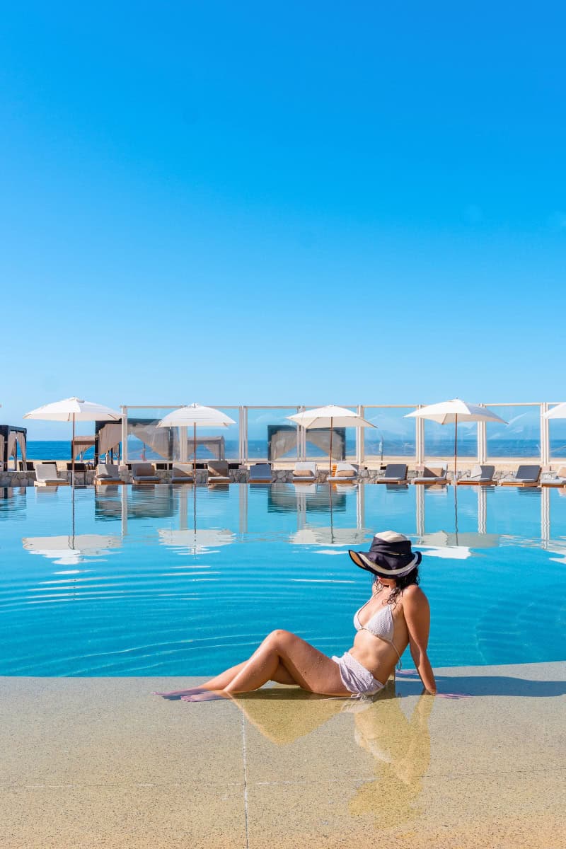 Relaxing woman in a swimsuit sitting by the poolside with ocean view and umbrellas in the background.